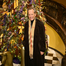 Richard E. Grant wearing a Burberry Scarf stood in front of a large Christmas tree covered in red, purple and mustard bows.