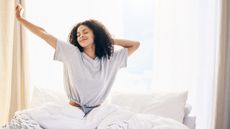 How to get up earlier image shows a woman with curly black hair stretches and smiles as she wakes up early on a sunny summer's morning
