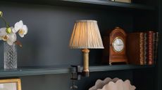 A bookshelf lamp with a gold base and neutral pleated shade on a dark blue-painted bookcase with books, a clock and decorative objects 