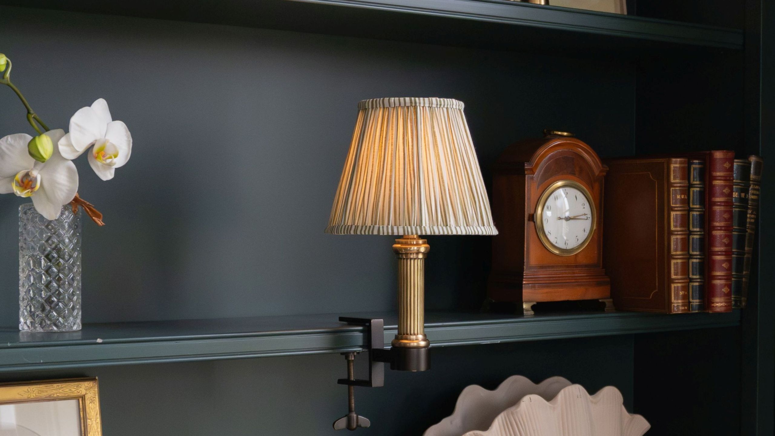 A bookshelf lamp with a gold base and neutral pleated shade on a dark blue-painted bookcase with books, a clock and decorative objects 