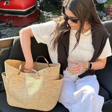 British Anna Newton poses on a boat while holding a drink and looking through her straw tote bag wearing black sunglasses, a linen ivory t-shirt, brown sweater over her shoulders, and white jeans