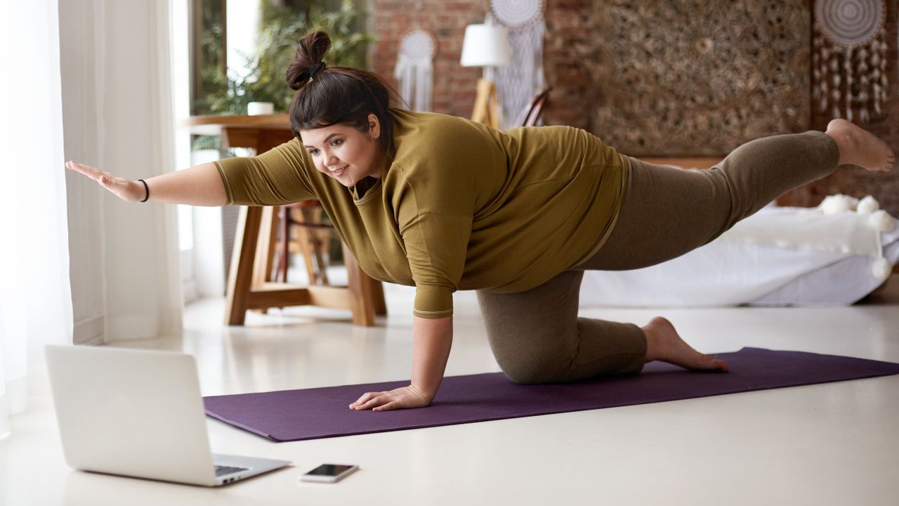 A woman in a long-sleeved top and leggings performs a bird-dog move in a lbedroom on an exercise mat. She is on her hands and knees, with her right arm off the ground and pointing forward, and her left leg elevated and pointing backward. Behind her we see a bed, lamp and desk.