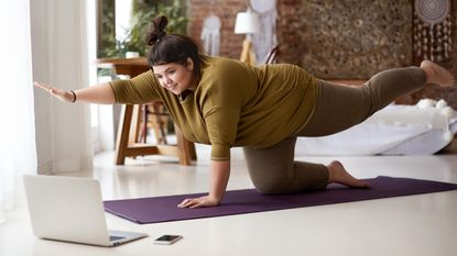 A woman in a long-sleeved top and leggings performs a bird-dog move in a lbedroom on an exercise mat. She is on her hands and knees, with her right arm off the ground and pointing forward, and her left leg elevated and pointing backward. Behind her we see a bed, lamp and desk.