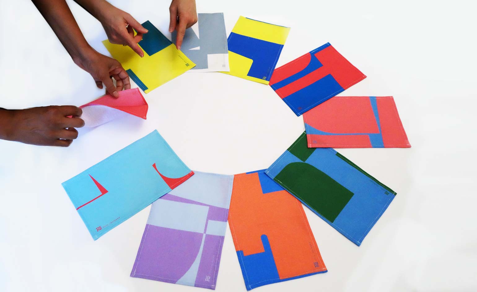 The hands of 2 individual laying out ten abstract cloth table napkins on a white surface
