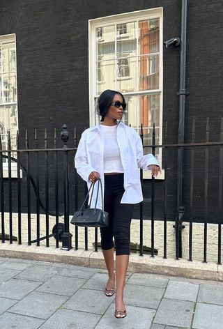 a photo showing the best summer-to-fall transition items with a woman walking down the street wearing a white button-down shirt over a white tank top styled with black capri pants, a black shoulder bag, and black mules