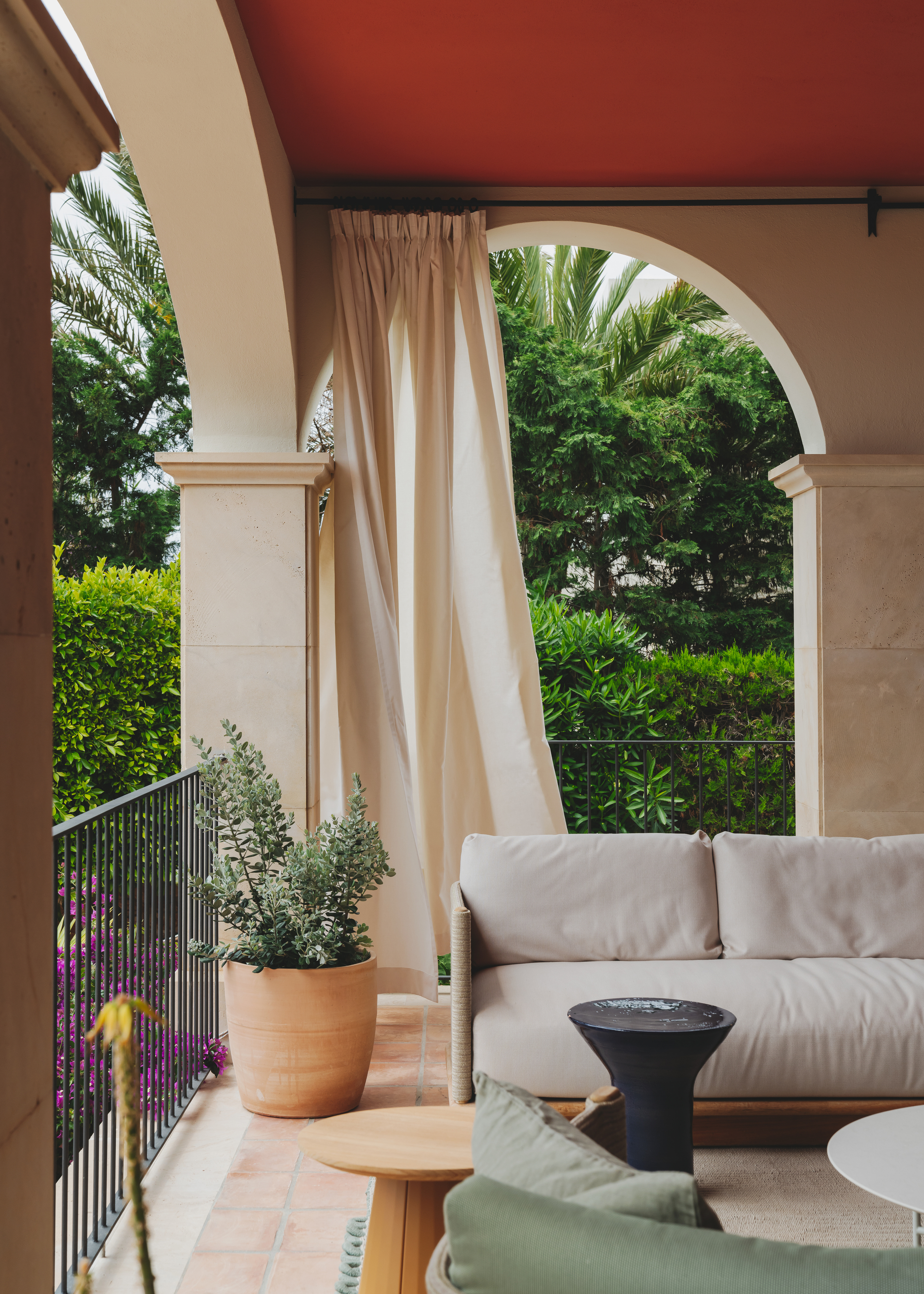 An outdoor terrace area with garden furniture, a ceiling painted red, and beige curtains hanging between the arches
