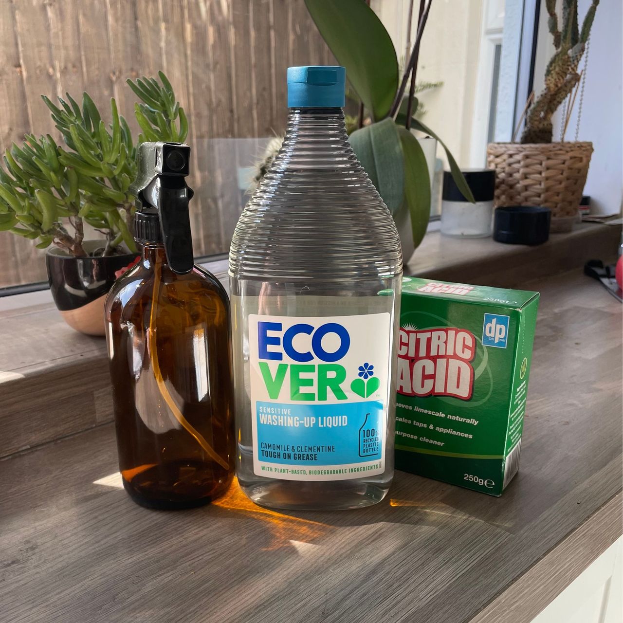 A brown glass bottle with spray top, a bottle of ecover washing up liquid, and a box of citric acid sitting next to each other on kitchen worktop