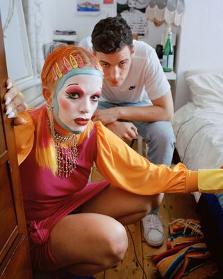 A close-up image of two people. Front: A person with short orange hair, a clown face paint, orange dress and large earrings and necklace. Behind, a white male with a white Nike tee-shirt, denim trousers and white shoes.