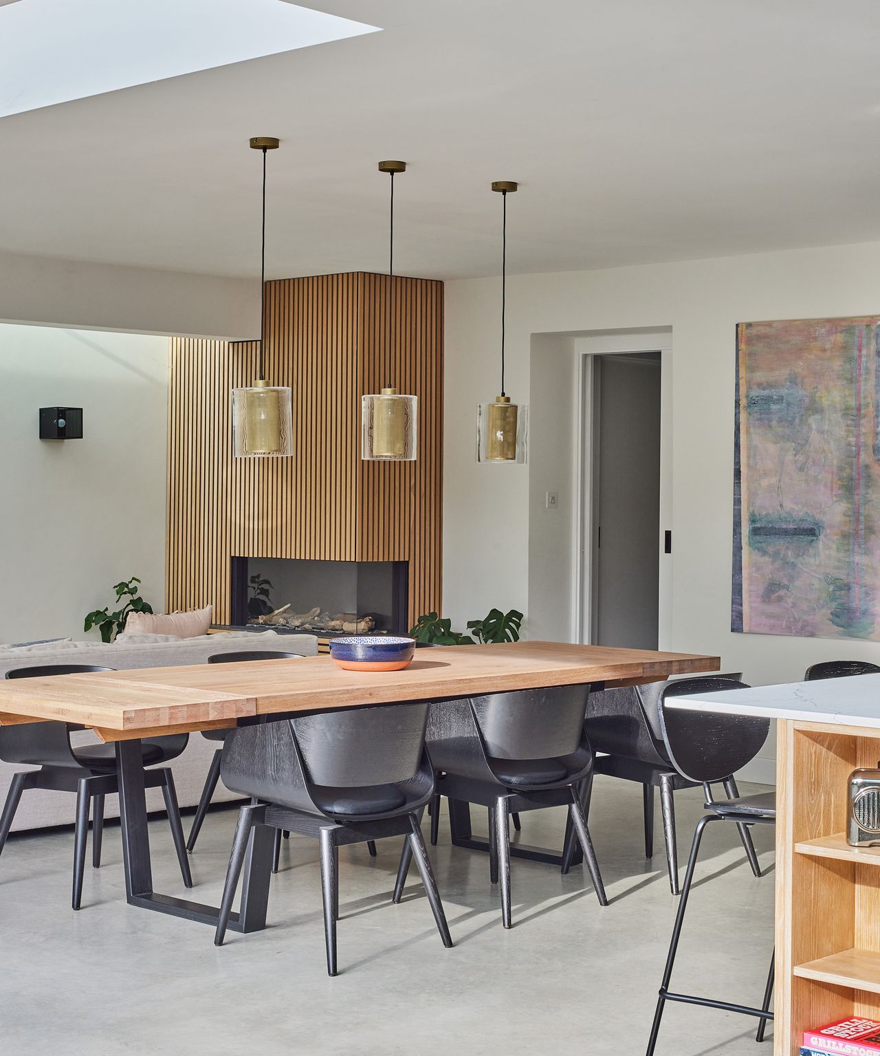 A large wood dining table with black metal legs and contemporary black chairs seated around. There are three gold pendant lights above the table