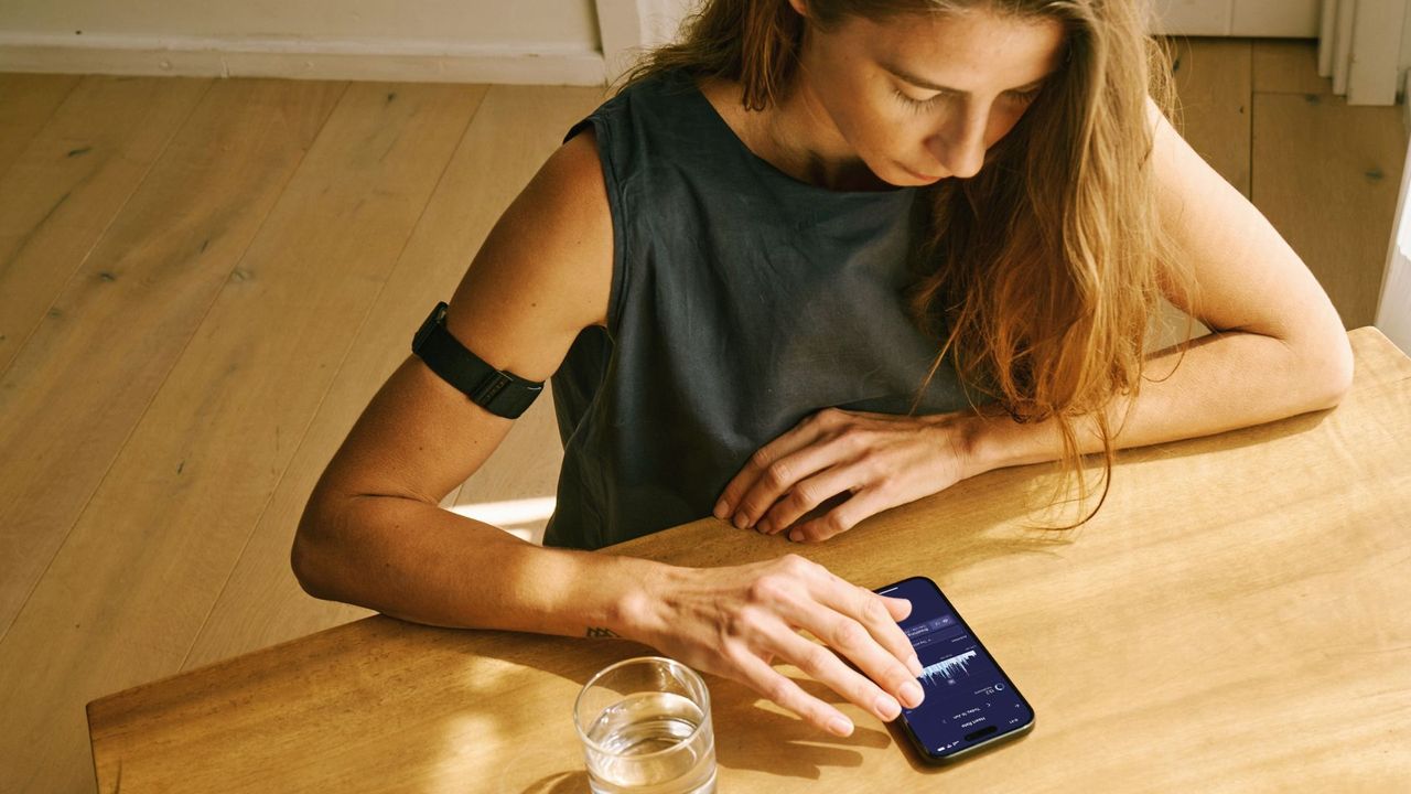 A woman wearing a Visible armband sits at a table looking at her phone. The Visible app is just visible behind her fingers