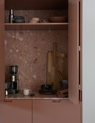 Image of a dark, muddy pink painted cabinet that opens into a coffee bar station. The countertops and backsplash of the bar are a pink terrazzo material. There are items like a coffee maker, mugs, and cutting boards.