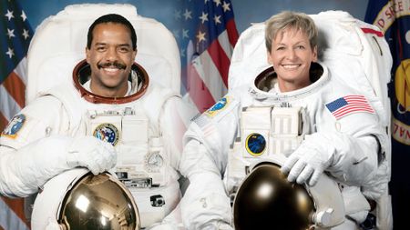 a Black male and caucasian female astronaut in white spacesuits are seen in portraits taken on the ground 