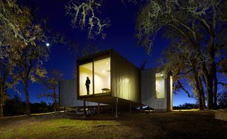 Exterior view of Moose Road House with the lights on and surrounding greenery at night. There is a person inside standing by the window and the moon can be seen in the distance