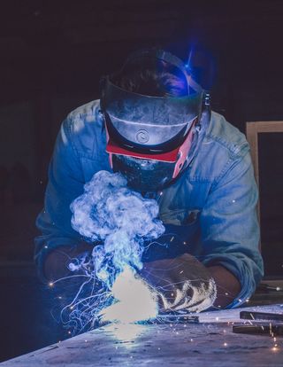 View of Mats Christéen in a denim shirt, welding helmet and gloves welding material