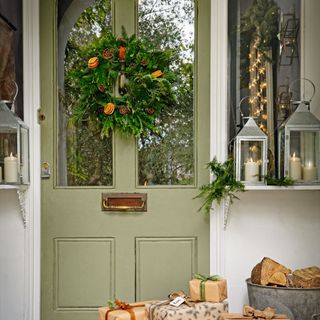 Pale green front door with a Christmas wreath, lanterns with candles either side, a sled on the doorstep with parcels wrapped in brown papers and ribbons