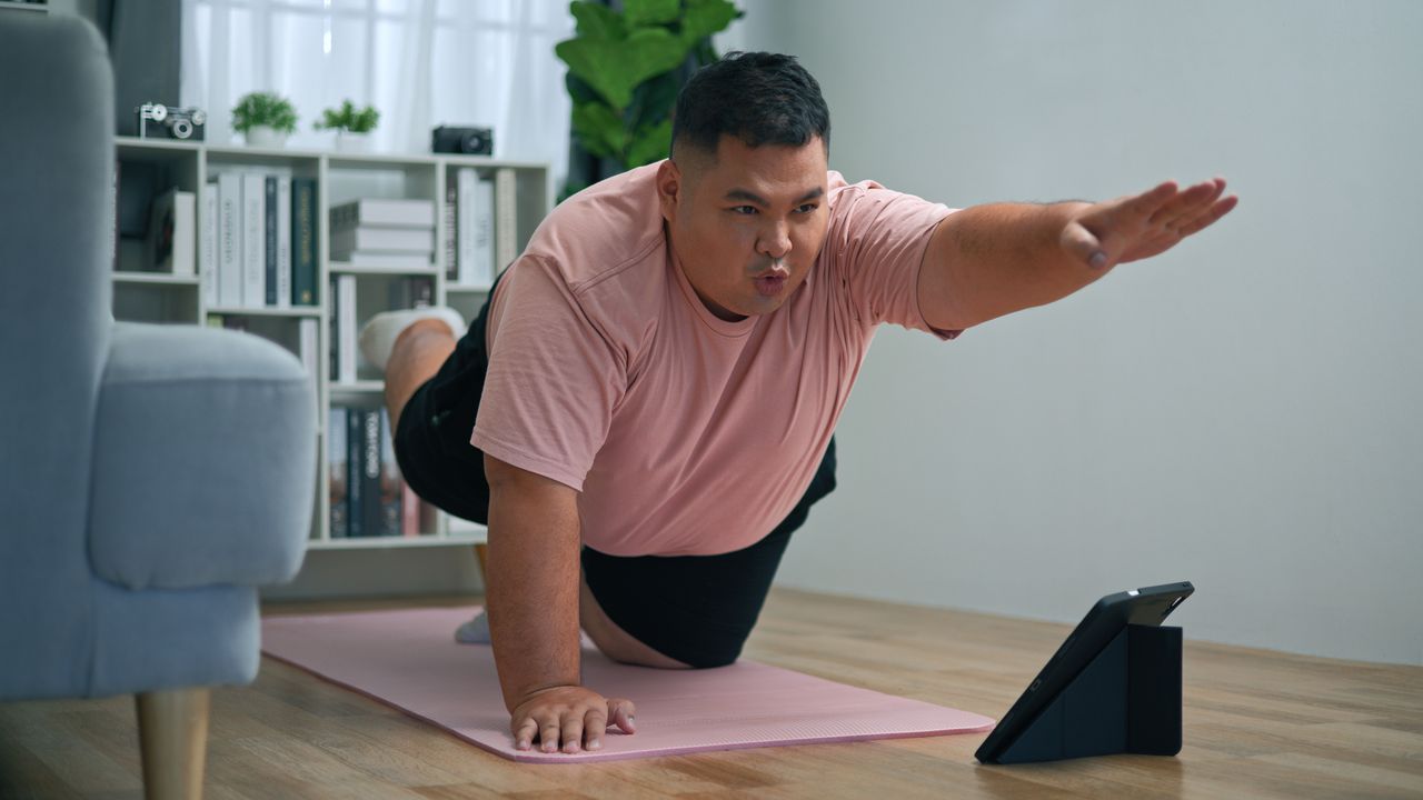 A man performs the bird dog exercise in a living room on a yoga mat. From all-fours, on his hands and knees, he is lifting his left arm up and forward and right leg up and backward, so that they are parallel with the floor. His other hand and leg are on the floor supporting him. We see a chair and shelving nearby.