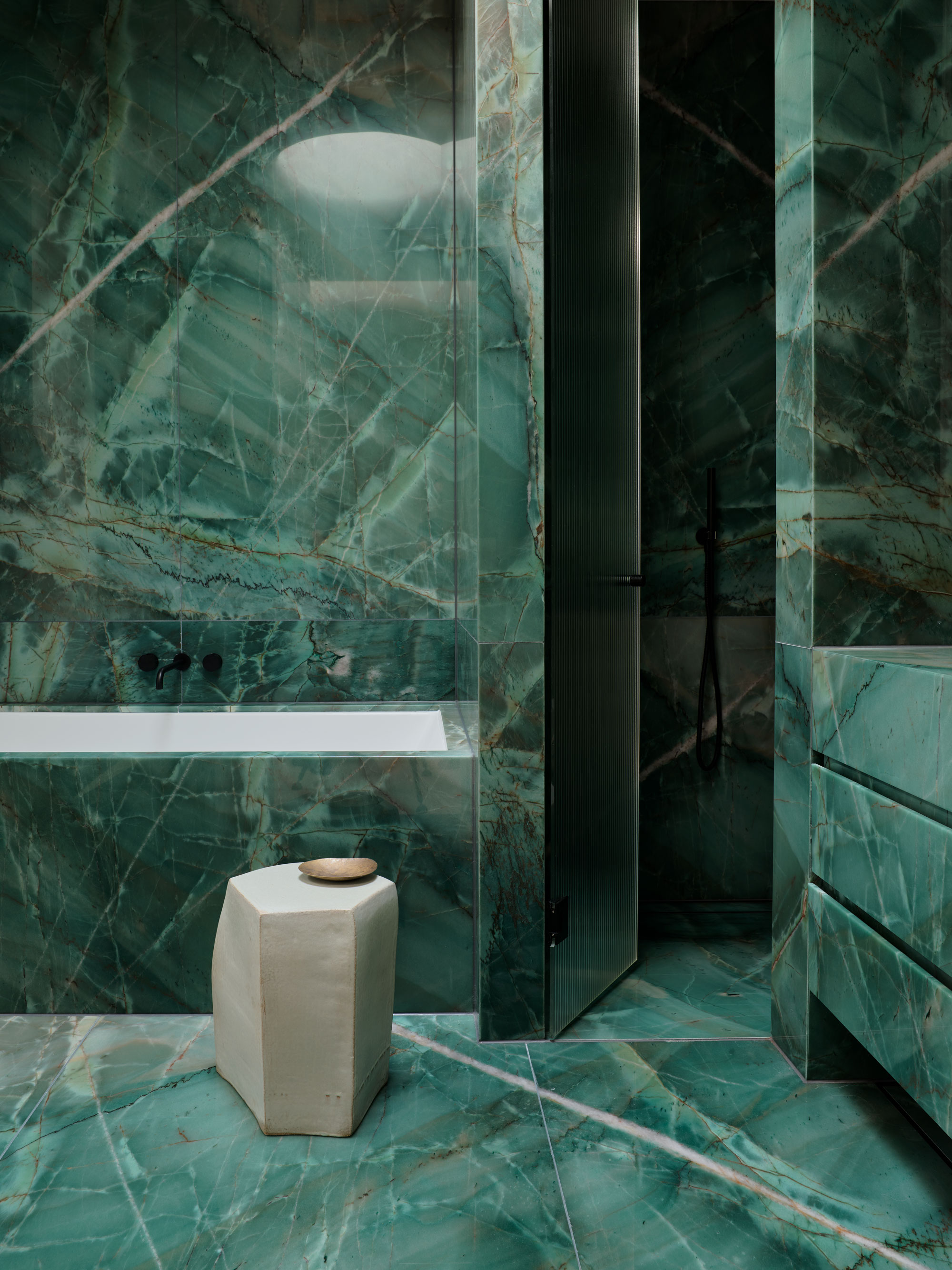 An emerald green, tone covered bathroom with a view of the built in bath tub and a small stool next to it and a set of drawers to the right.