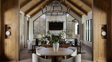 Open-concept rustic home interior featuring a round dining table in the foreground, leading into a high-ceilinged living room with large geometric windows and vaulted wood beams.