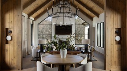 Open-concept rustic home interior featuring a round dining table in the foreground, leading into a high-ceilinged living room with large geometric windows and vaulted wood beams.