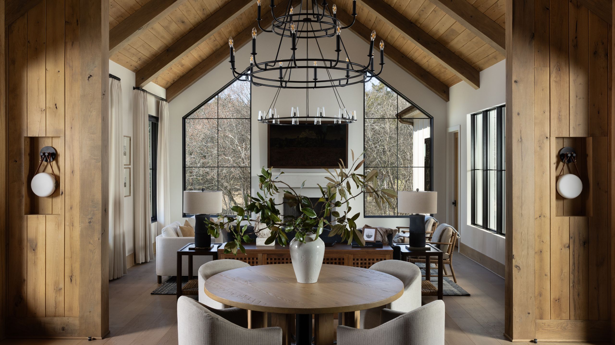 Open-concept rustic home interior featuring a round dining table in the foreground, leading into a high-ceilinged living room with large geometric windows and vaulted wood beams.