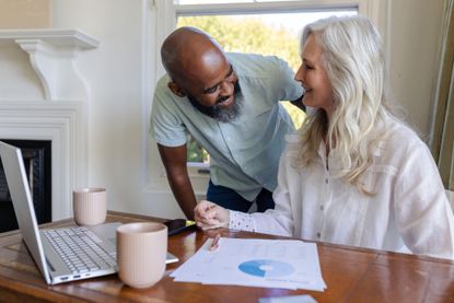 3CHMYKB Couple smiling at home, discussing wedding plans with laptop and coffee mugs