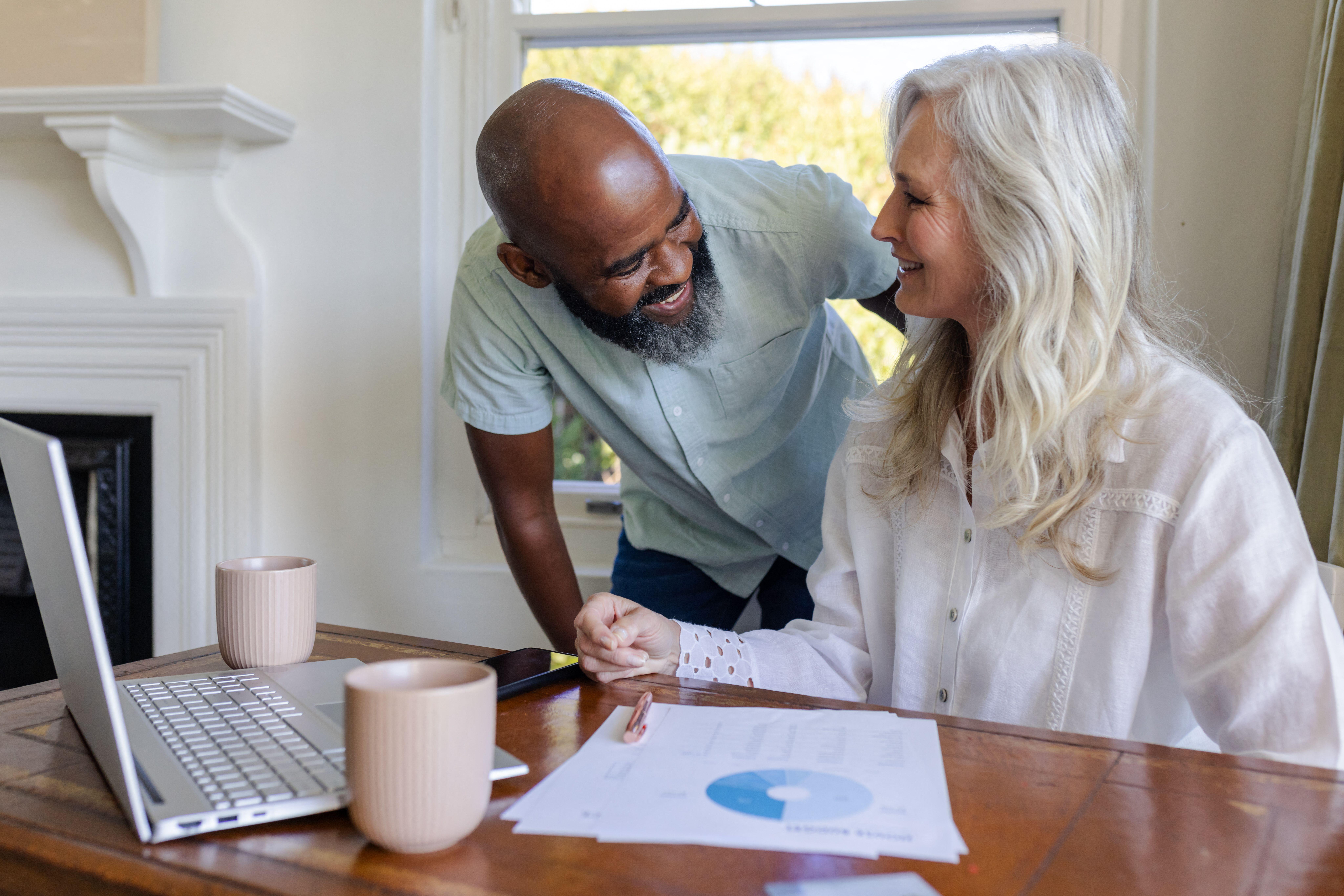 3CHMYKB Couple smiling at home, discussing wedding plans with laptop and coffee mugs