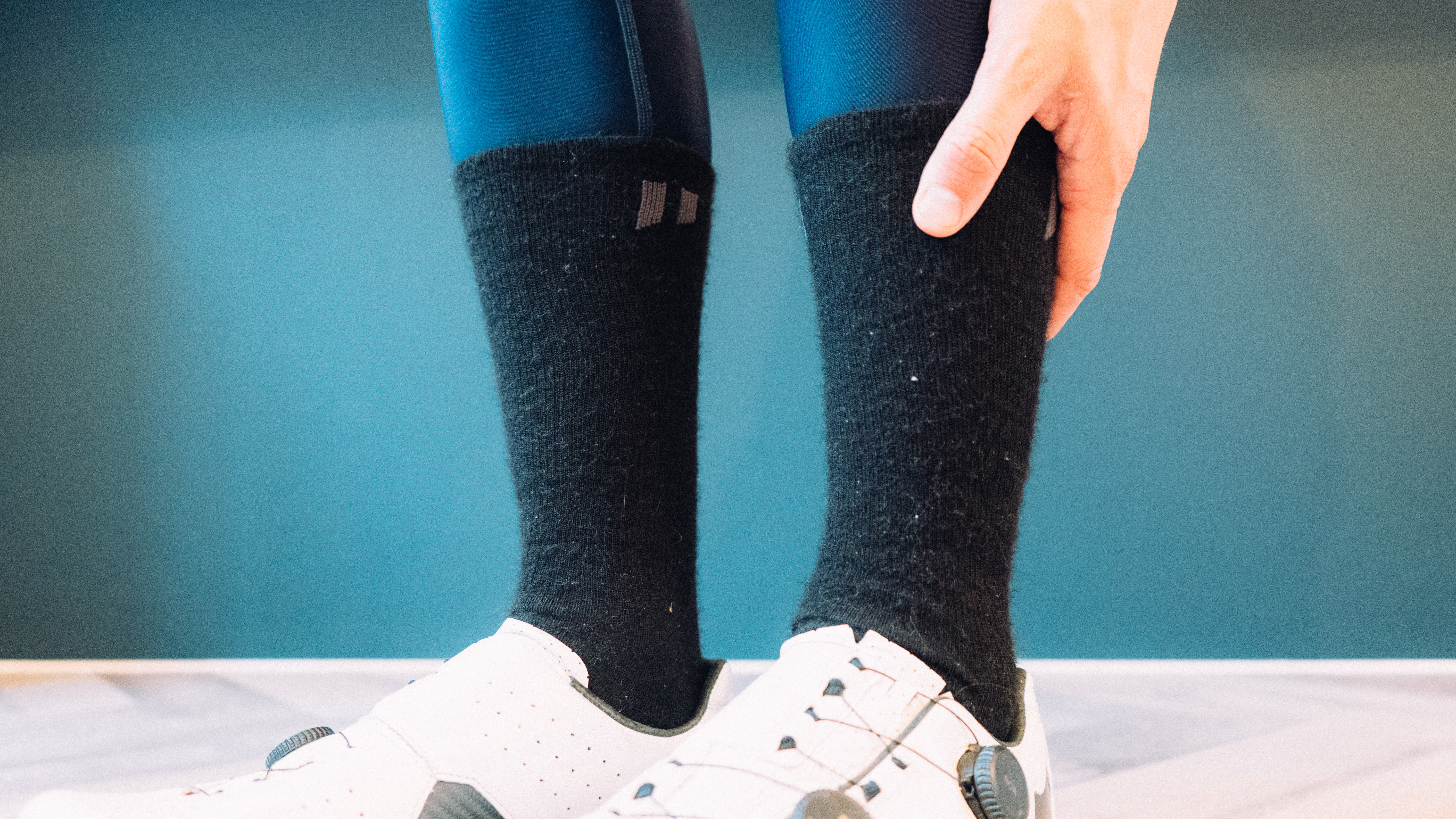 A man wears a set of white cycling shoes, black fluffy socks, and blue tights against a blue background