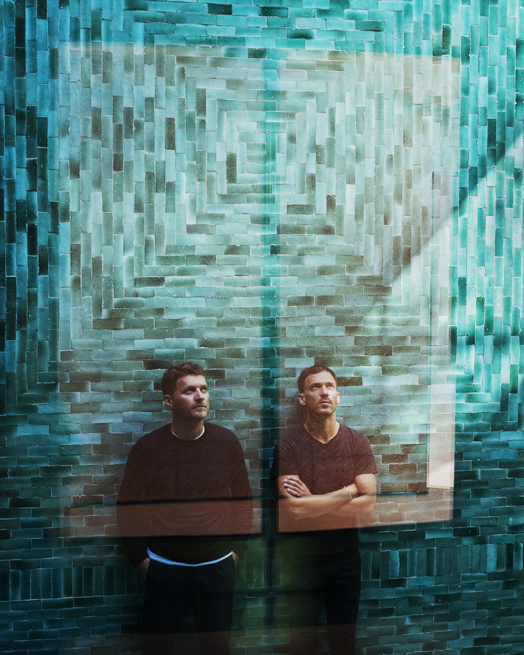 Studio KO founders portrait, two men looking upwards standing against a blue tile wall
