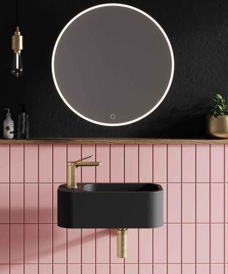 A small bathroom with a black wall-mounted sink with a gold faucet, light pink splashback tiles, and a black wall above it with a circular mirror above it