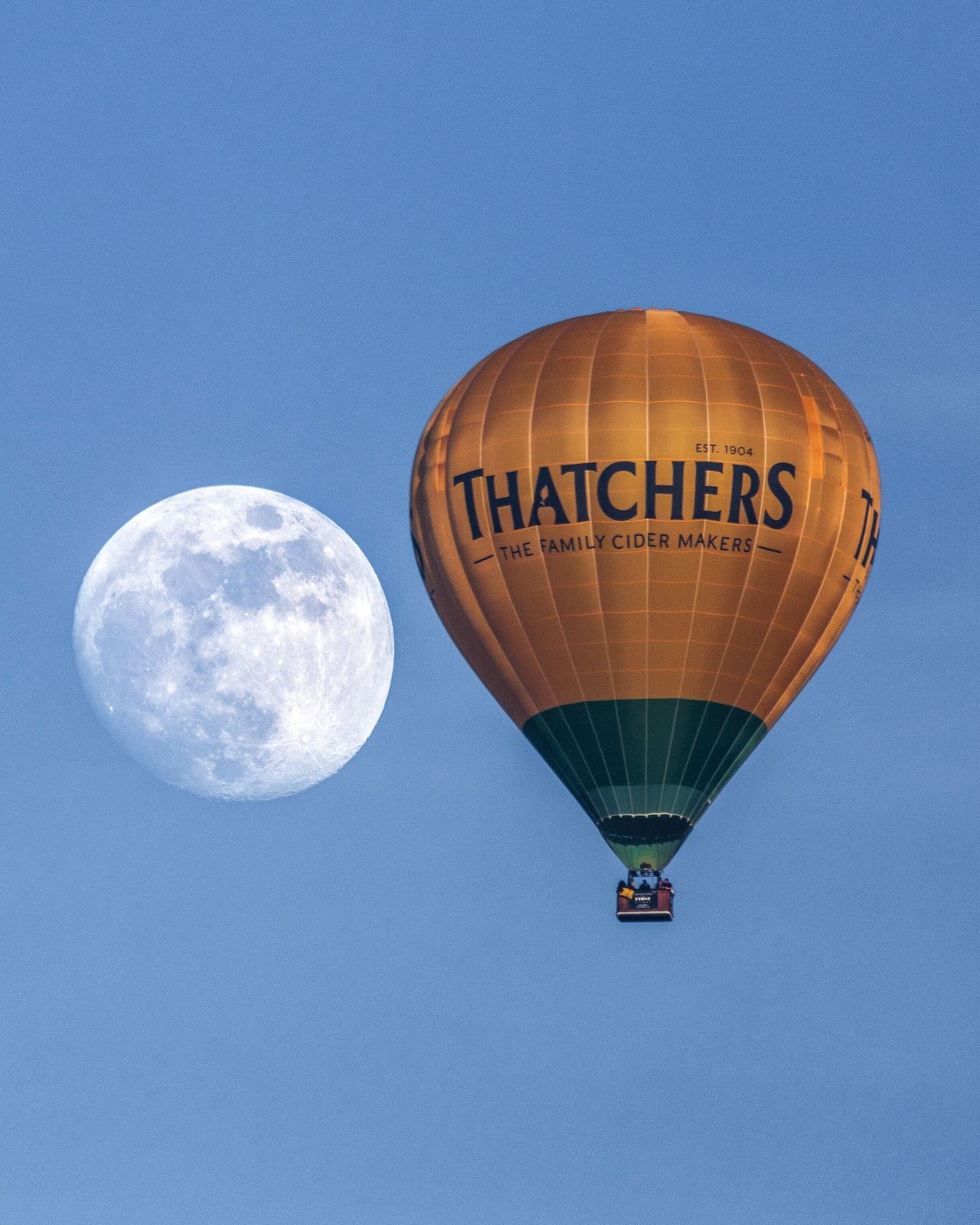 a fully illuminated moon to the left of a hot air balloon against a blue sky.
