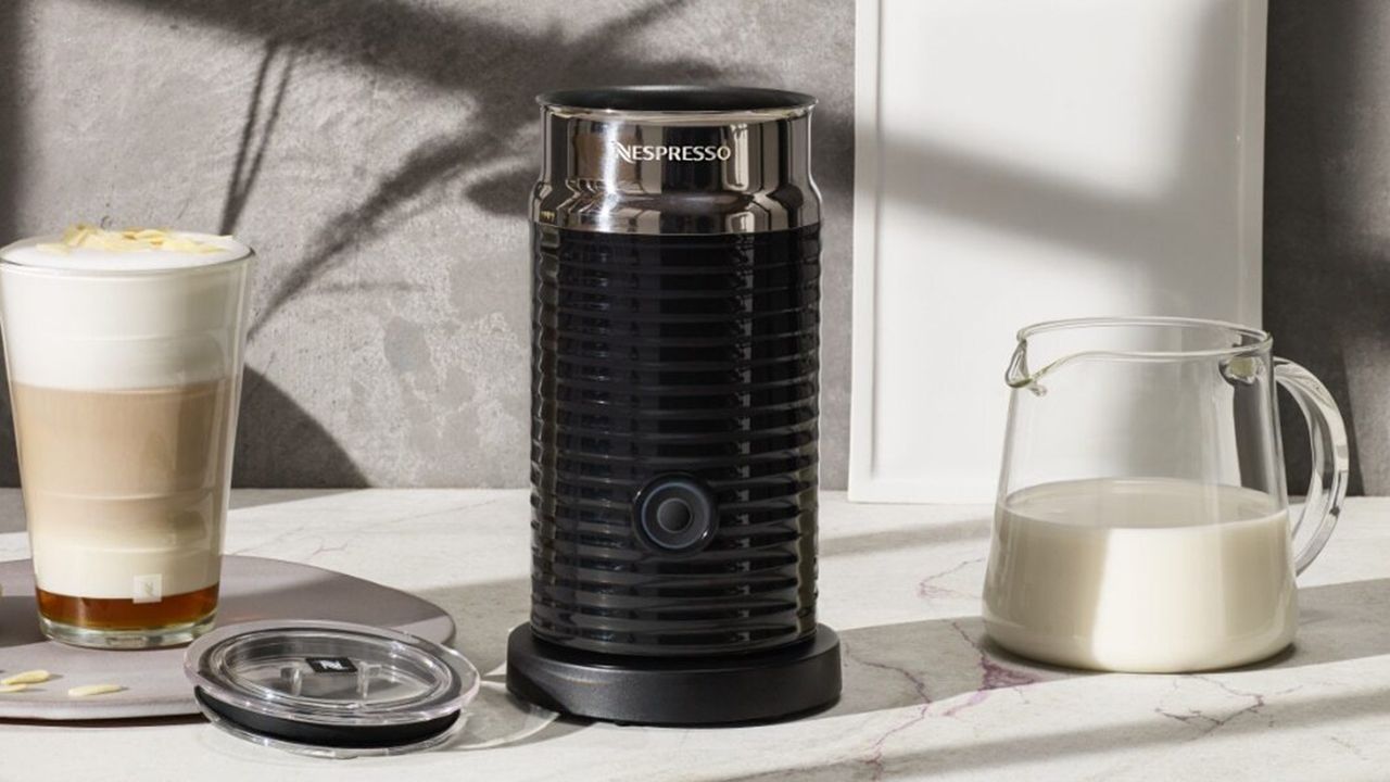 Black Nespresso Aeroccino 3 with glass jug containing cows&#039; milk and prepared coffee-based beverage on marble countertop