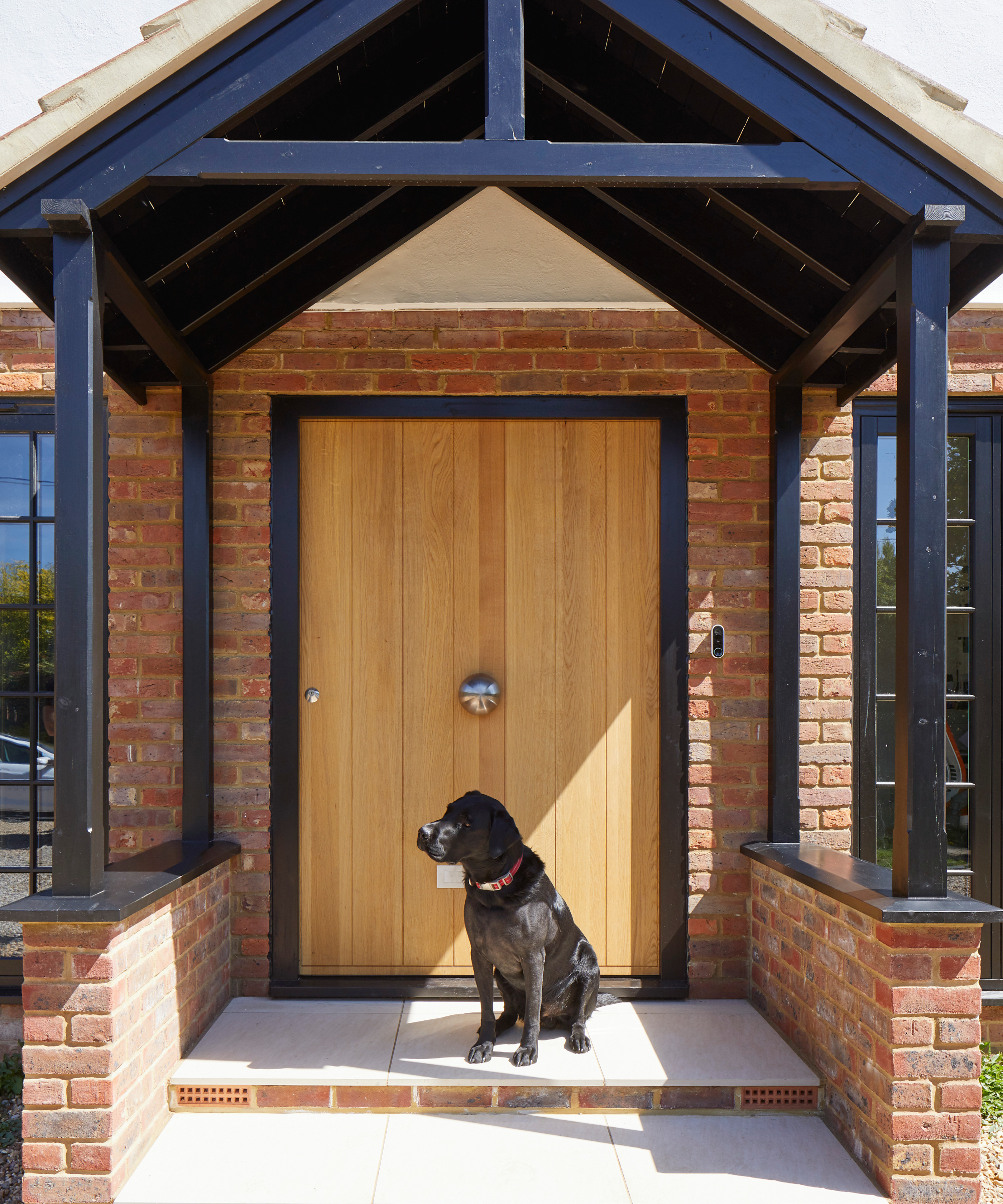 front porch with small brick dwarf walls, pitched roof, open sides, black painted framework leading to oak front door with black frame, black labrador sat on steps