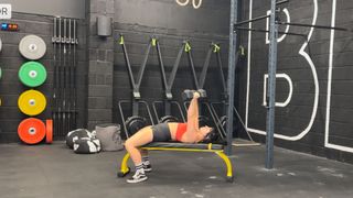 Raquel Sanjurjo performs a dumbbell chest press in a gym. She is lying flat on a weights bench, feet on the floor, and holding a pair of dumbbells directly above her head.