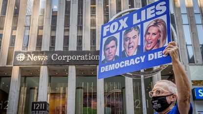 man holds up sign that reads "Fox lies, democracy dies" with the faces of Fox News correspondents on it outside the news stations' corporation