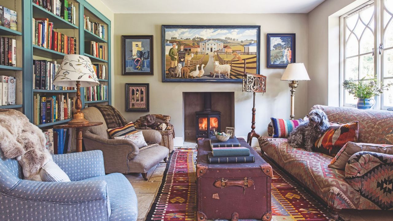 Cozy cottage living room with sofa, two armchairs and chest placed upon a rug, surrounding a fireplace with gallery wall and bookshelves