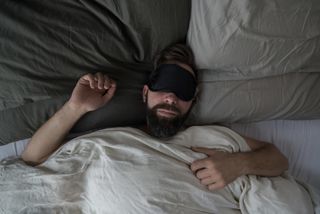 A man wearing a black sleeping mask while laying in bed.