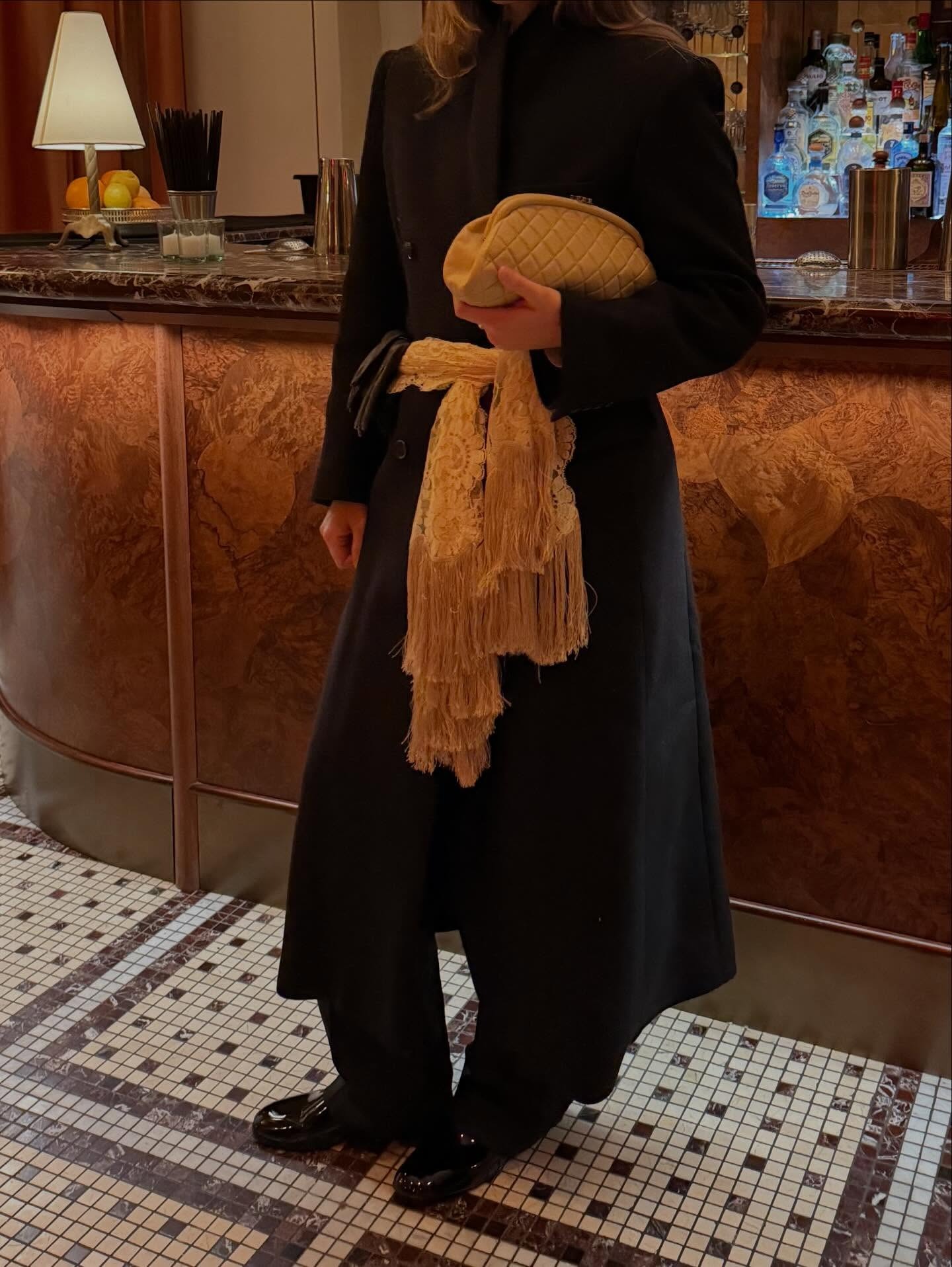 @anna__laplaca wearing a black coat, cream clutch, and cream lace scarf with fringe tied around her waist.