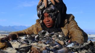 A woman wearing a fur coat and a black headscarf adorned with mussels and a starfish lies on a sandy beach behind a pile of seashells.