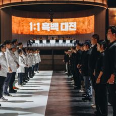 Two rows of chefs (the White Spoons, left, and the Black Spoons, right) stand facing each other, with a circular dining table in the back and a screen reading "1:1" with a Korean phrase, in 'Culinary Class Wars.'