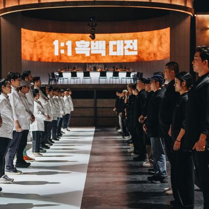 Two rows of chefs (the White Spoons, left, and the Black Spoons, right) stand facing each other, with a circular dining table in the back and a screen reading "1:1" with a Korean phrase, in 'Culinary Class Wars.'