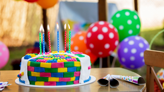 Colorful, LEGO-inspired birthday cake with 6 multicolored candles sits on a table with three noisemakers. There are polka-dotted birthday balloons in rainbow colors in the background.