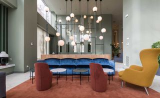 A hotel sitting area with, two burgundy chairs, a yellow chair, two round coffee tables, a shiny blue sofa, an orange rug and artistically shaped round pendant lights