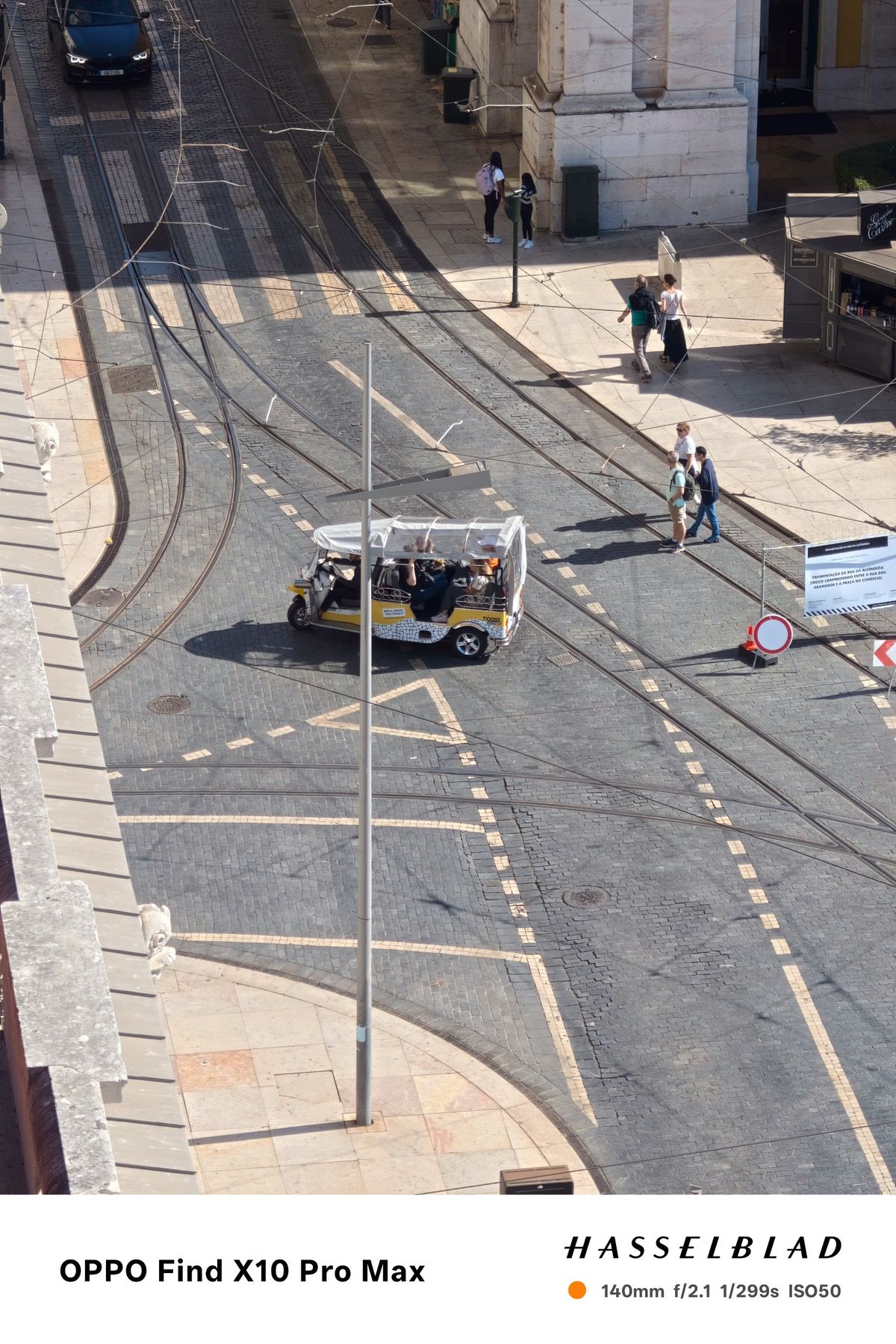 Tuk-tuk driving across tram tracks on a sunlit Lisbon street viewed from above.
