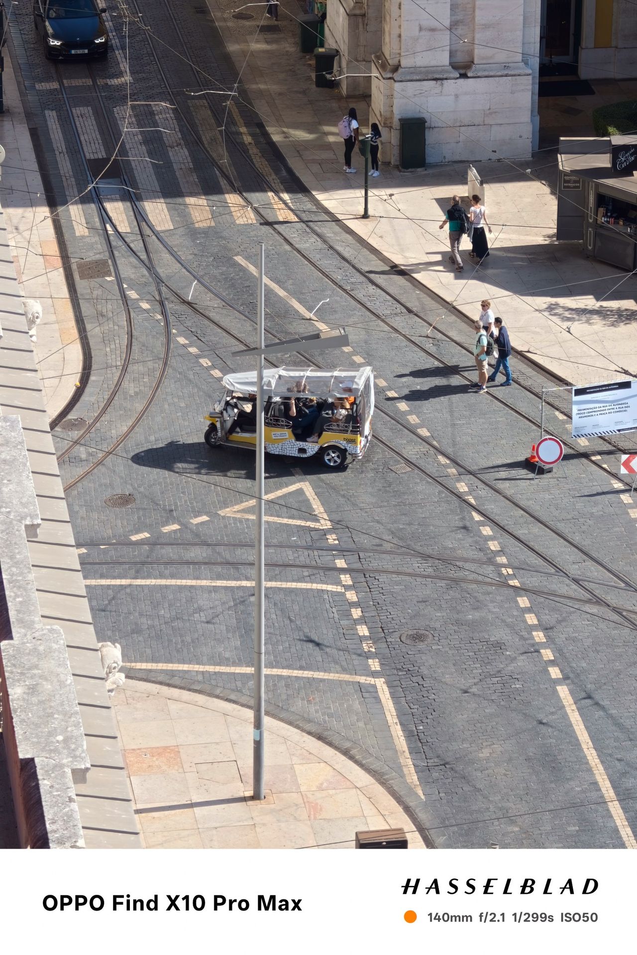 Tuk-tuk driving across tram tracks on a sunlit Lisbon street viewed from above.