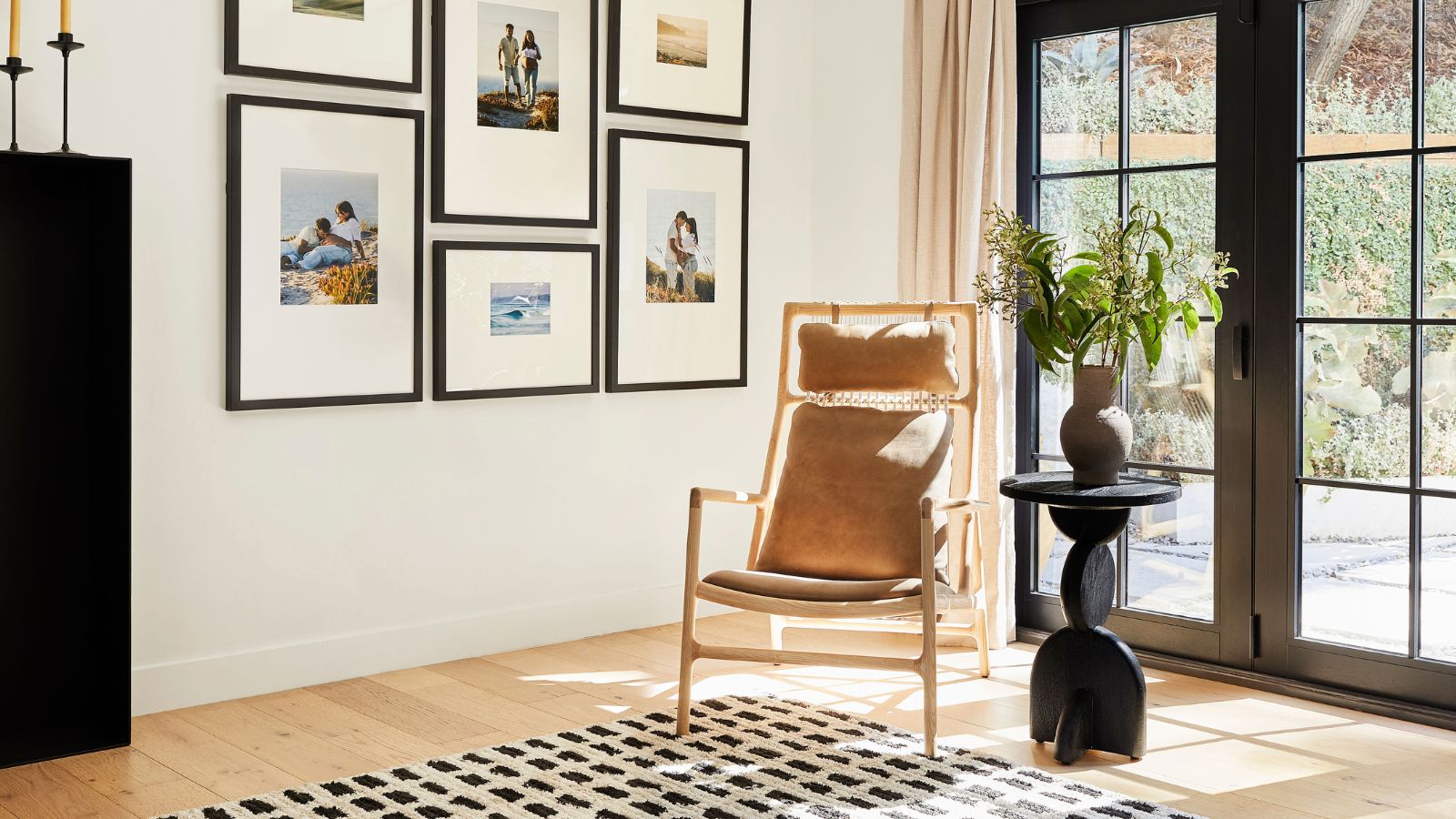 Bright room corner with gallery of framed photographs, rattan and wooden armchair, side table with plant and vase, and large, paneled windows revealing bright outdoor area.