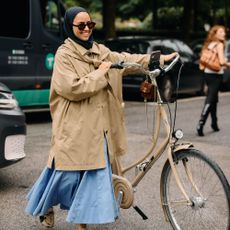 Copenhagan Fashion Week attendee wears an oversized trench coat with a baby blue voluminous skirt. She pushed a bicycle wears wears cow print shoes.