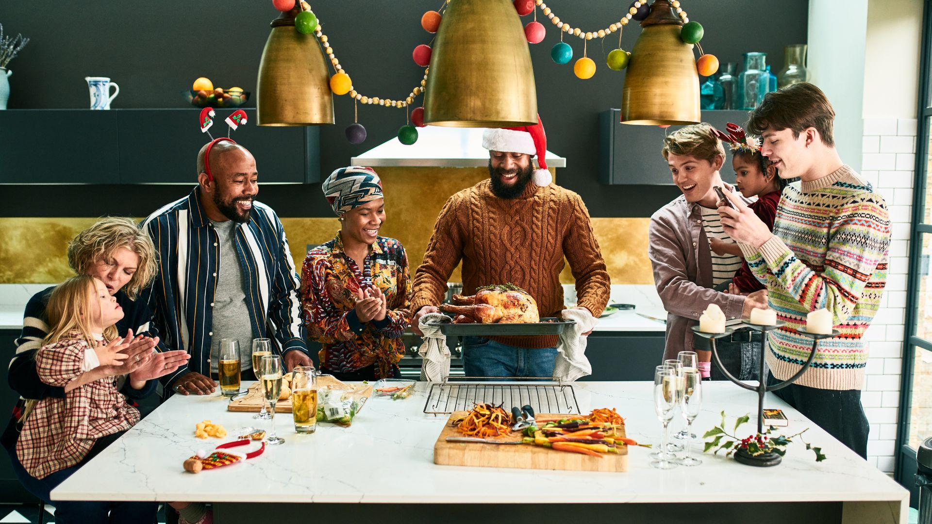 A group of smiling people standing around a table at Christmas. In the center, a man in a Santa hat holds a tray with a turkey on it.
