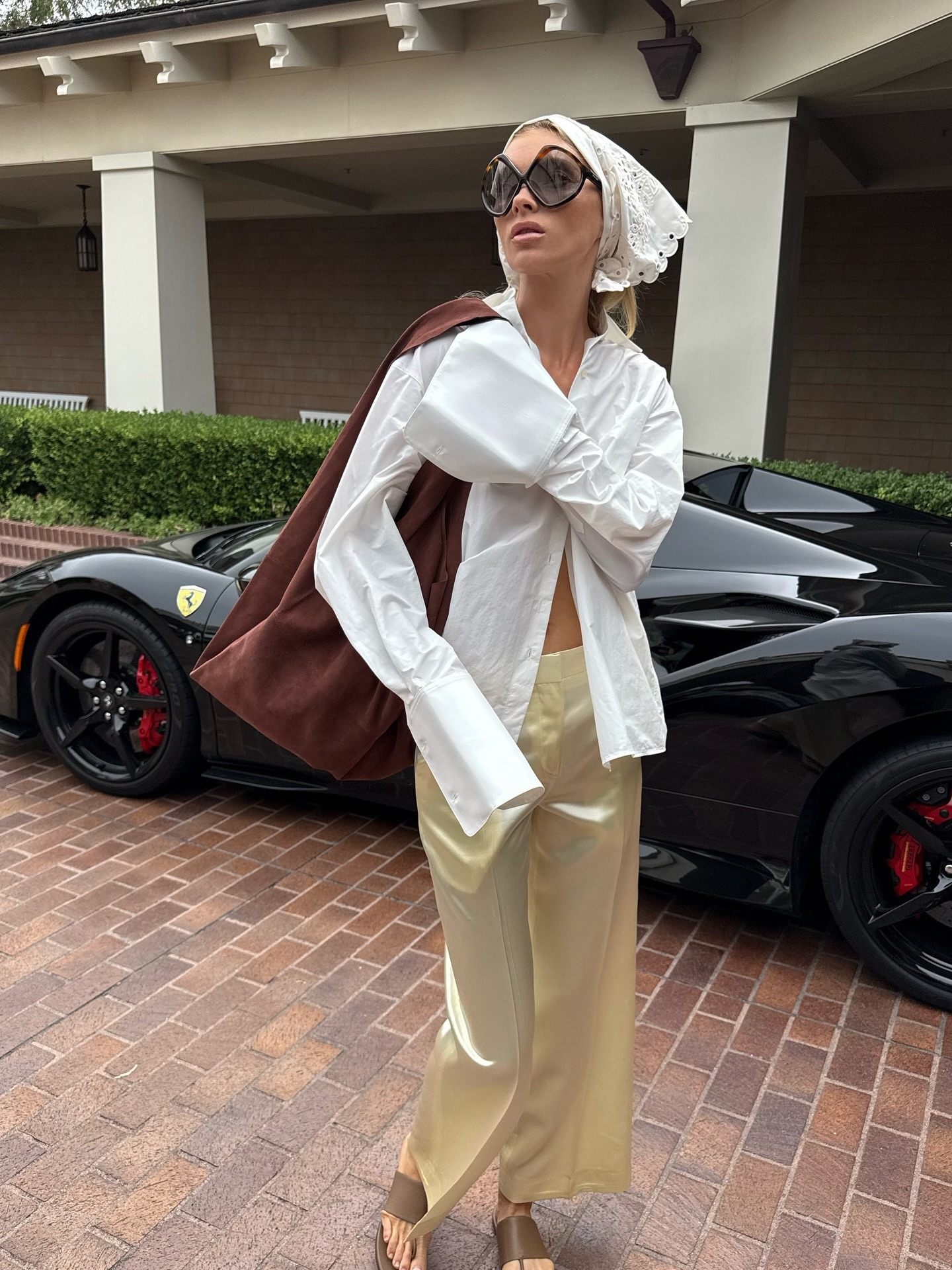 Elsa wears a white bandana on her head, a brown suede bag, a white button-down, ivory silk pants, and tan sandals.
