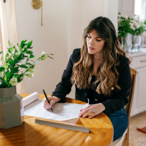 Image of a woman in a black jacket and jeans sitting at a wooden table drawing on a piece of architecture paper.