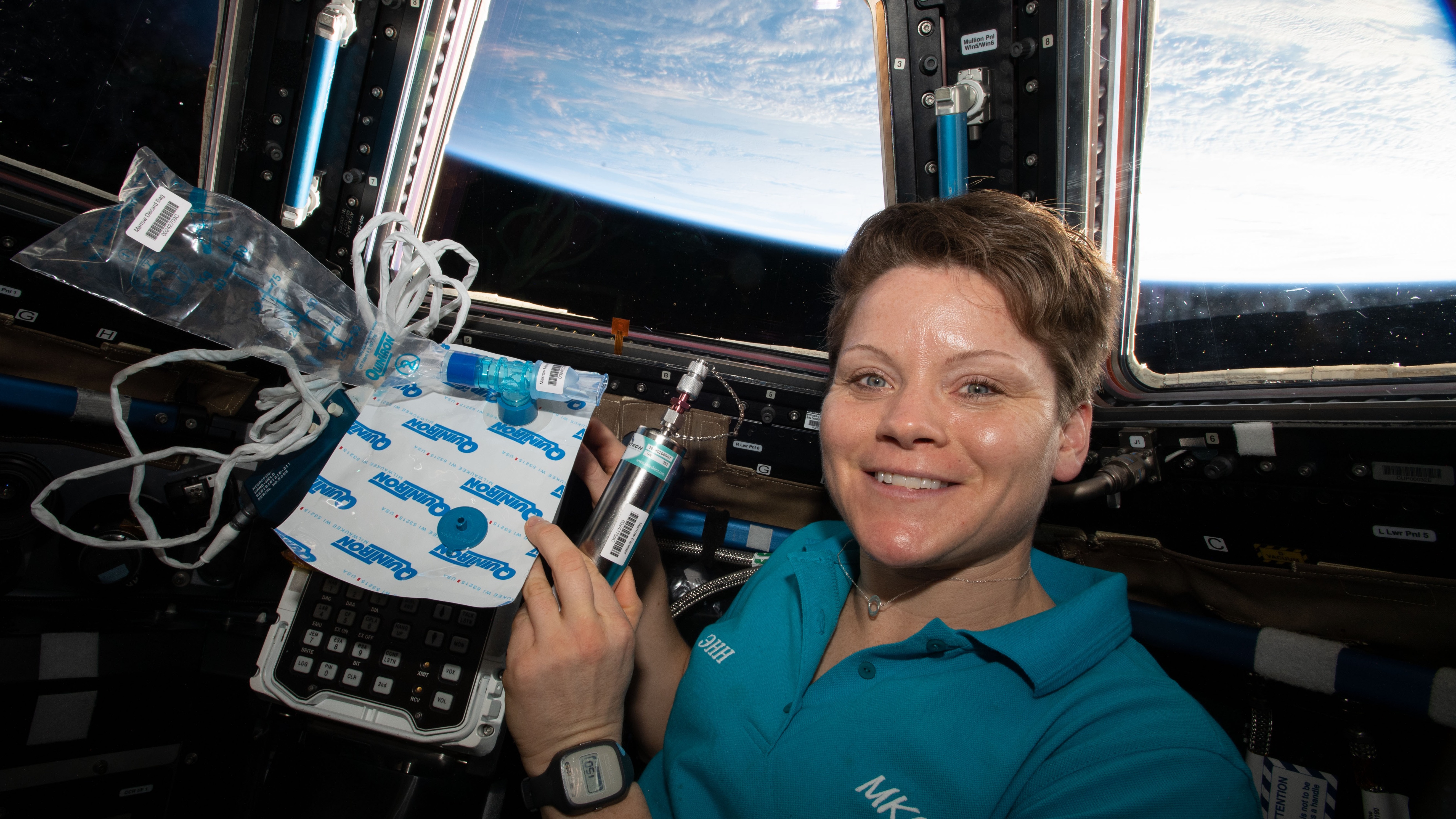 a woman with short hair poses for a picture aboard the international space station, with earth visible through windows behind her